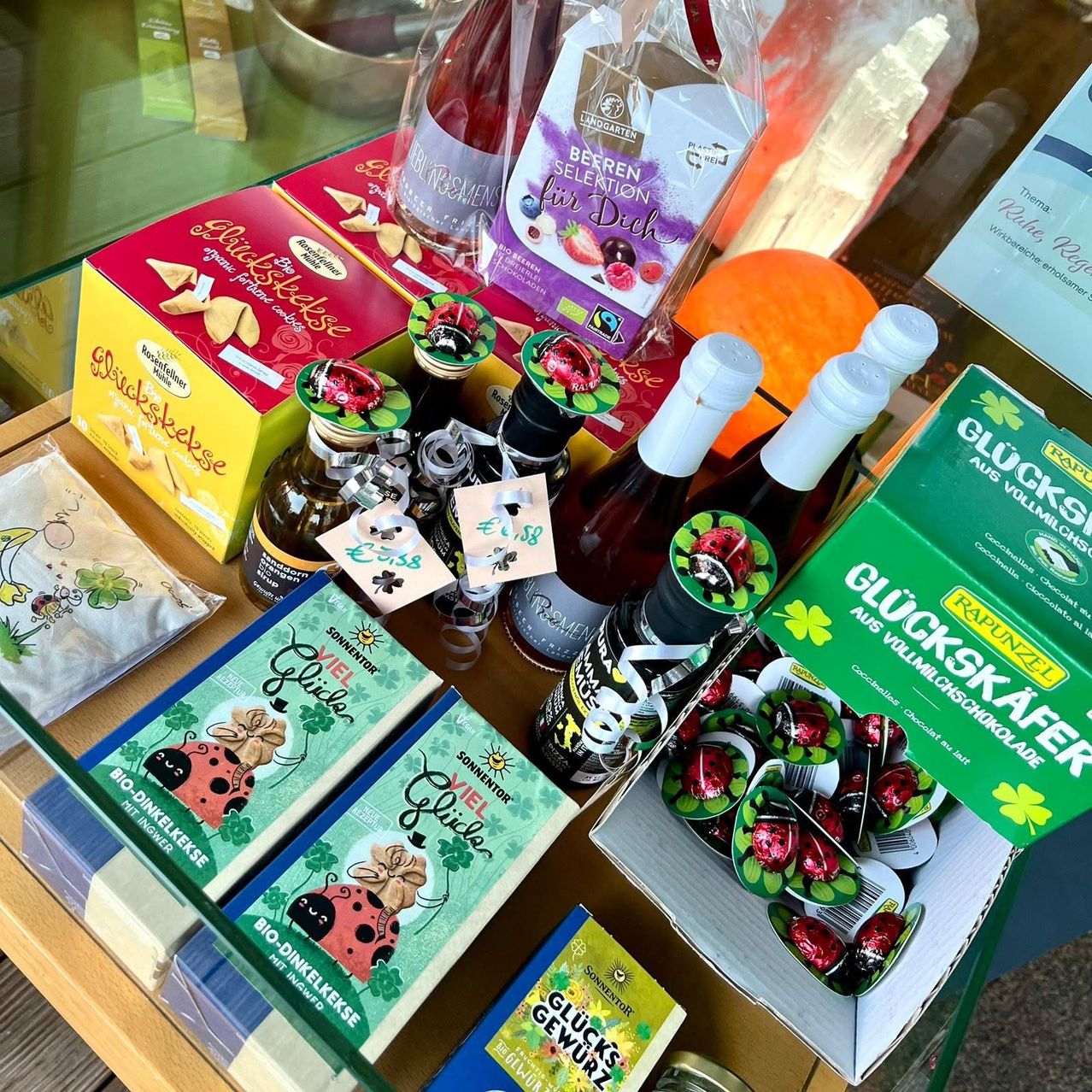 Ein Holztisch präsentiert verschiedene Lebensmittelprodukte. Eine rote Schachtel mit der Aufschrift 'Glückskekse' ist sichtbar. Flaschen mit grünen Kappen und Etiketten befinden sich daneben. Andere Schachteln tragen die Bezeichnungen 'BIO-Dinkelkekse', 'Glückskäfer' und 'Glücksrevierz'. Die Artikel sind ordentlich angeordnet, möglicherweise in einem Geschäft.