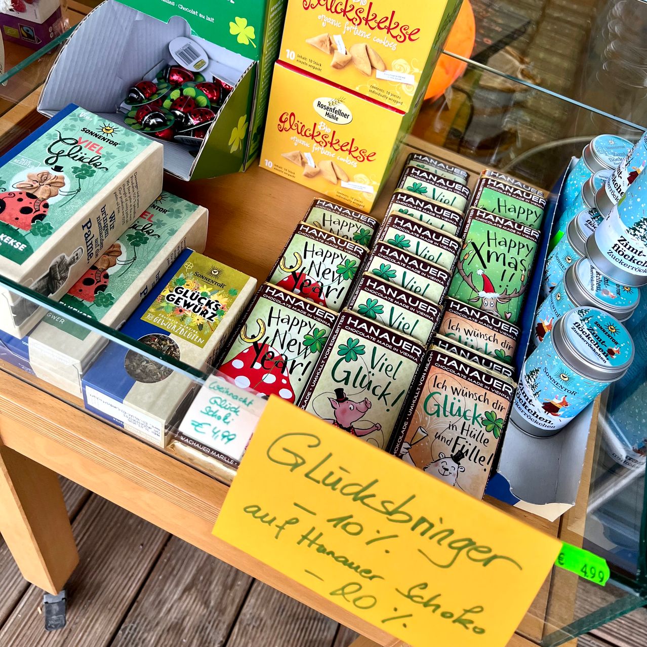 Ein Holzregal präsentiert verschiedene Produkte wie Schokoladen, Tees und Süßigkeiten. In der Mitte befindet sich ein gelbes Schild mit der Aufschrift 'Gluecksbringer ab 10! 20% Rabatt'. Verschiedene farbenfrohe Schachteln und Dosen sind angeordnet. Der Hintergrund zeigt eine Glaswand mit Reflexionen.