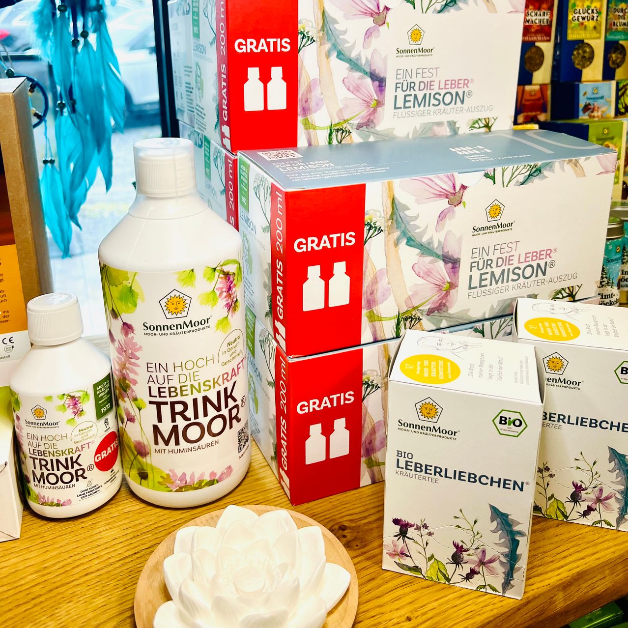 Auf einem Holztisch sind verschiedene Produkte ausgestellt. Flaschen mit 'SonnenMoor'-Etiketten, eine mit 'GRATIS', stehen neben Kartons mit pflanzlichen Bildern. Die größte Schachtel ist mit 'Eine feste für die Leber Limon' beschriftet. Andere Schachteln haben 'BIO' und 'Leberleibechen'-Etiketten. Im Vordergrund befindet sich eine weiße Blume.