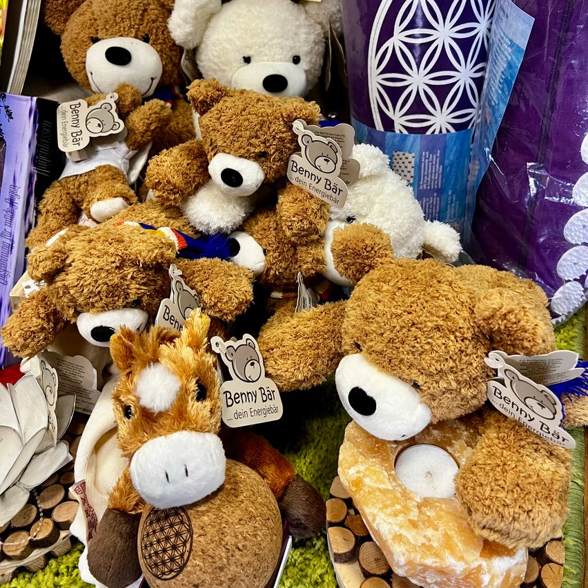 Eine Sammlung von Plüschtieren, darunter Bären und ein Pferd, auf einer grünen Oberfläche ausgestellt. Sie haben Etiketten mit Namen und Markeninformationen. Eine Kerze ist auf dem Bauch eines Bären platziert.