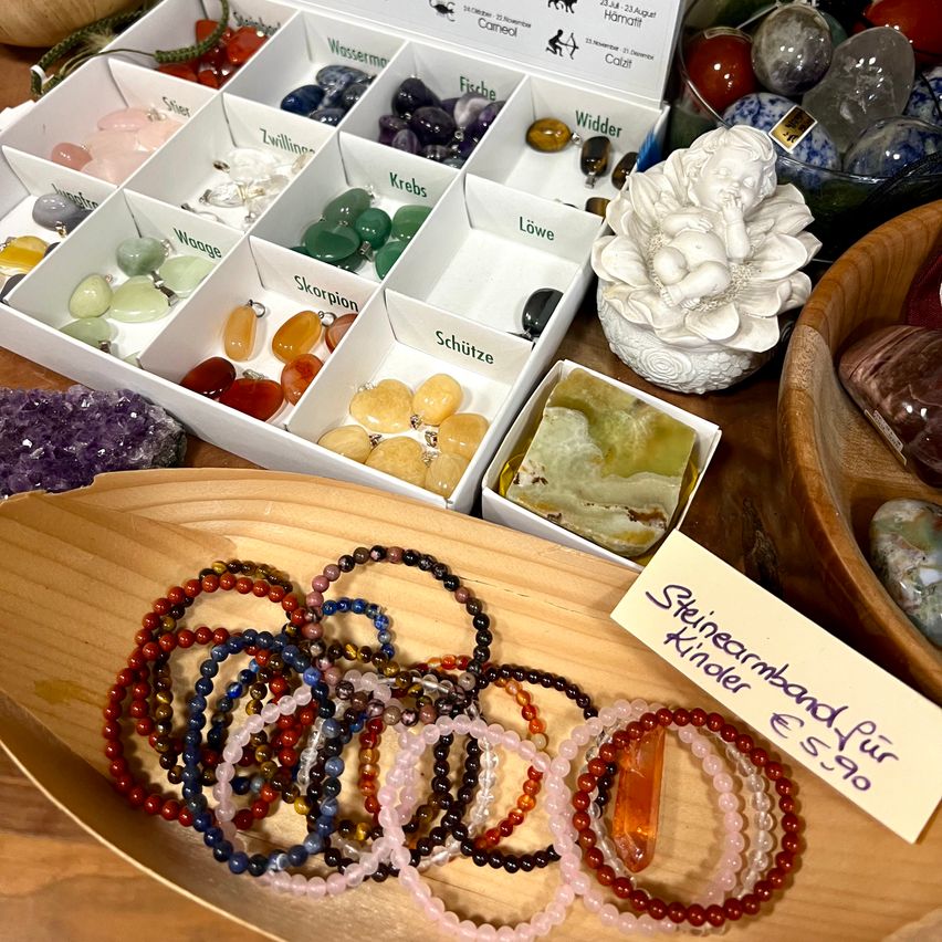 Ein Holztisch zeigt verschiedene Edelsteinarmbänder in einer Holzschüssel, verschiedene Kristalle in weißen Schachteln und eine Figur eines schlafenden Engels auf einer Blume.