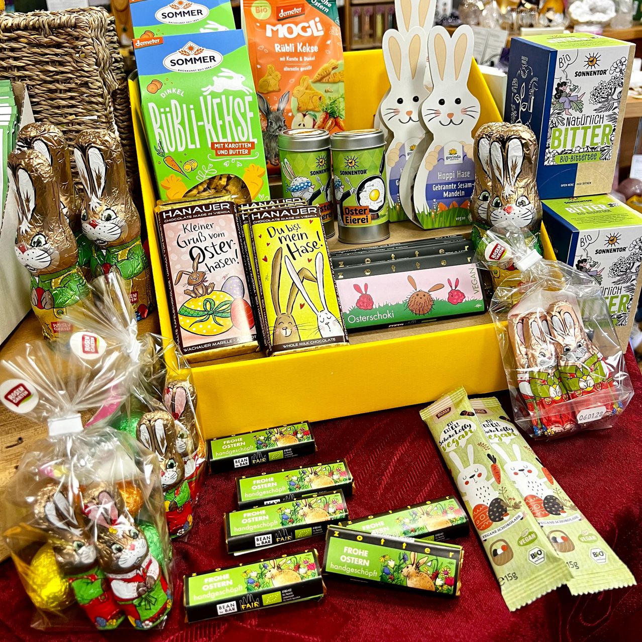 Eine gelbe Ausstellung mit verschiedenen Oster-Produkten, darunter Schokoladenhasen, Snacks und Süßigkeiten, auf einem roten Tisch. Es gibt auch Schachteln und Tüten mit Ostereiern und anderen festlichen Artikeln.