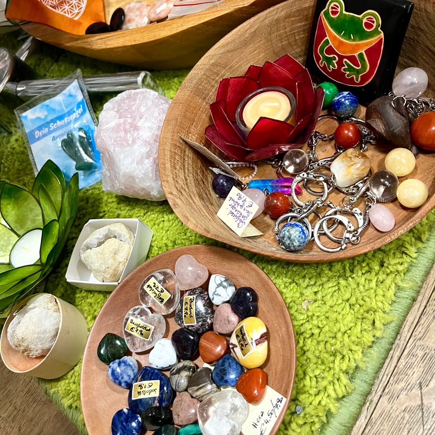 Eine hölzerne Schale mit verschiedenen Edelsteinen und Kristallen, einschließlich einer roten Lotuskerze, steht auf einer grünen Oberfläche. Eine weitere Schale mit einer Mischung aus Steinen befindet sich in der Nähe. Einige Gegenstände sind beschriftet, und links steht eine grüne Pflanze.