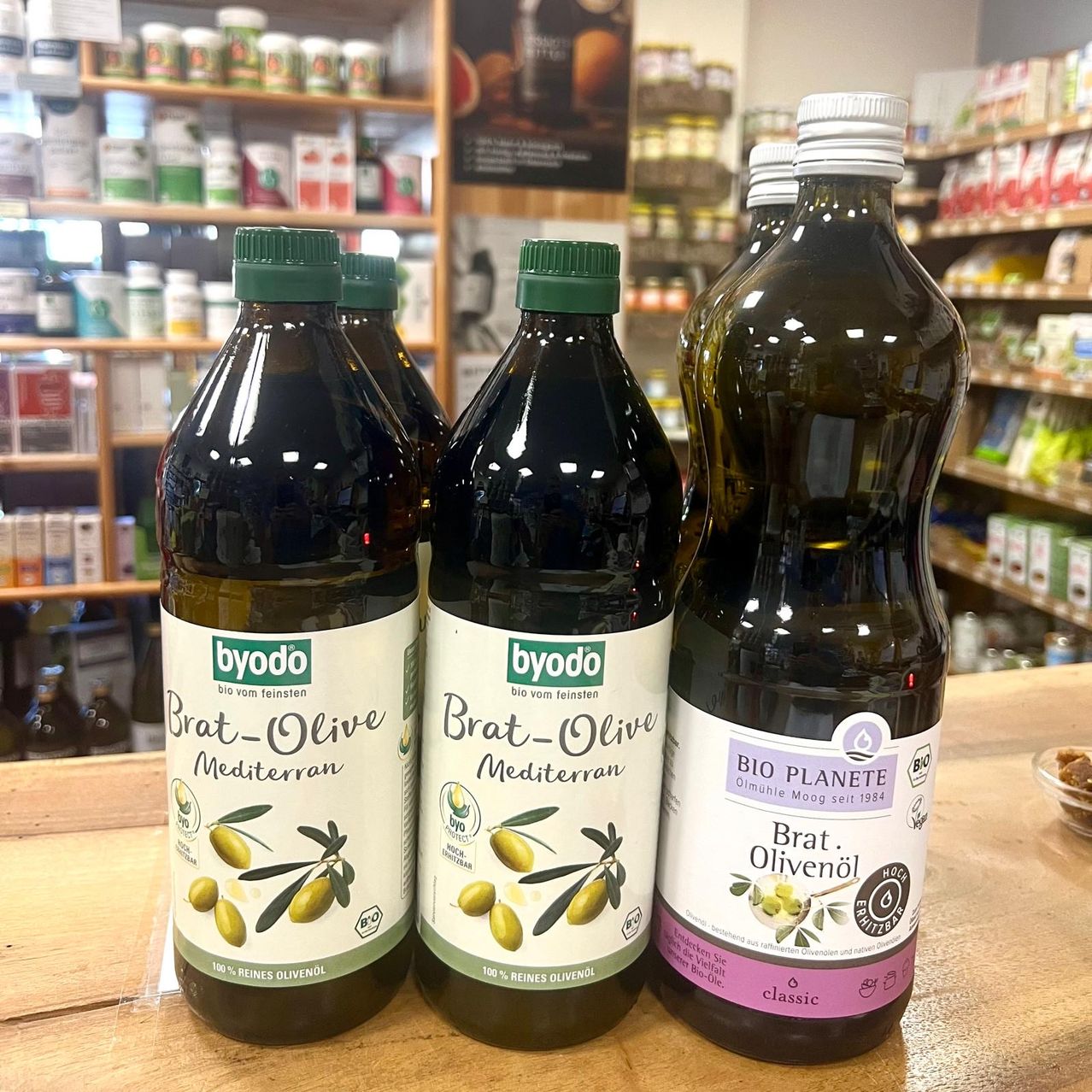 Drei Flaschen Olivenöl, zwei mit der Aufschrift 'byodo Brat-Olive Mediterran' und eine mit der Aufschrift 'BIO PLANETE Brat. Olivenol classic', sind auf einem Holztisch in einem Geschäft ausgestellt.