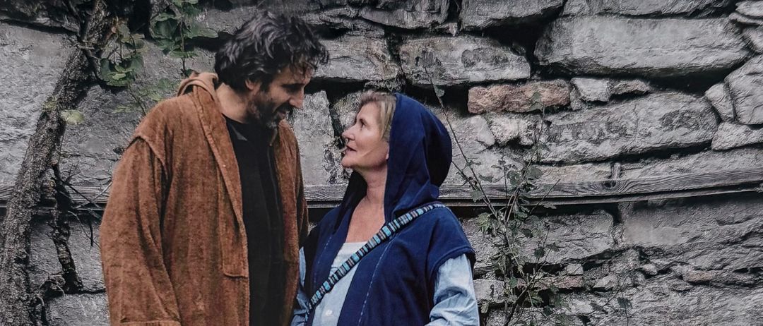 Ein erwachsener Mann und eine Frau stehen eng beieinander und teilen einen zärtlichen Moment. Sie sind beide in langen, blauen Gewändern gekleidet. Hinter ihnen steht eine Steinmauer mit Ranken und Pflanzen. Der Text lautet 'Anders als erwartet'.