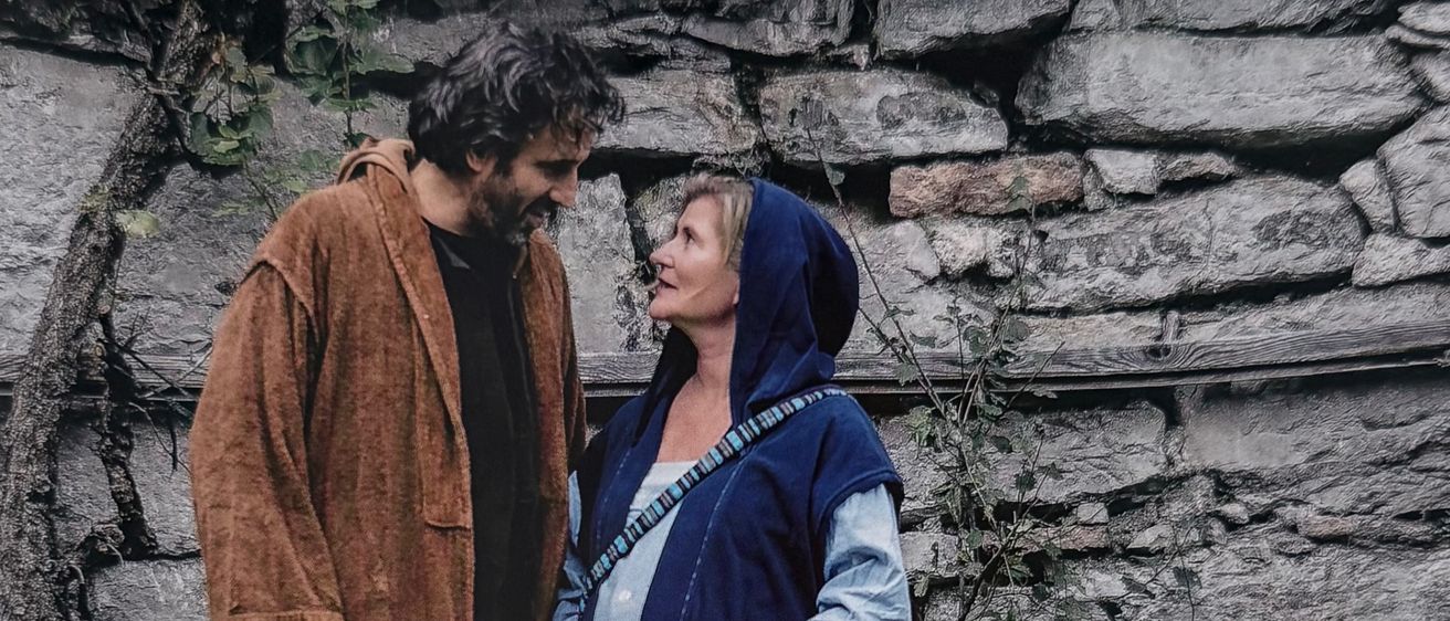 Ein erwachsener Mann und eine Frau stehen eng beieinander und teilen einen zärtlichen Moment. Sie sind beide in langen, blauen Gewändern gekleidet. Hinter ihnen steht eine Steinmauer mit Ranken und Pflanzen. Der Text lautet 'Anders als erwartet'.