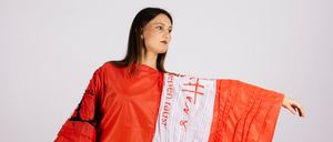 Eine Frau in einem roten, avantgardistischen Modekleid mit Text darauf steht vor einem weißen Hintergrund. Das Kleid zeichnet sich durch kühne Muster und eine dramatische Silhouette aus.