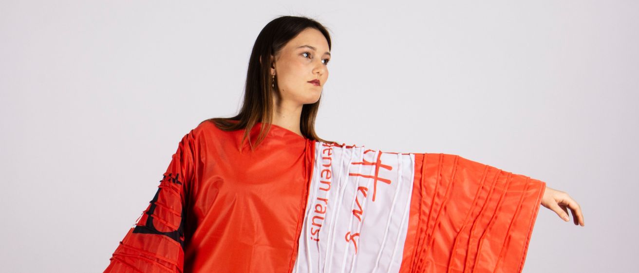 Eine Frau in einem roten, avantgardistischen Modekleid mit Text darauf steht vor einem weißen Hintergrund. Das Kleid zeichnet sich durch kühne Muster und eine dramatische Silhouette aus.