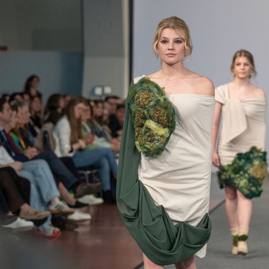 Eine Modenschau mit zwei Models, die die Laufstege hinuntergehen, eines trägt ein weißes Kleid mit grünem Stoff und das andere ein weißes Kleid. Zuschauer beobachten vom Sitzplatz aus.