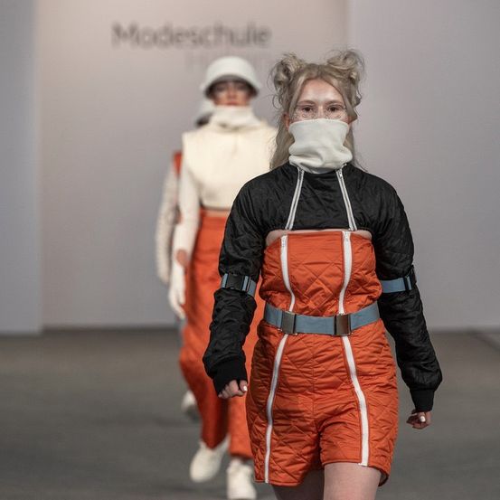 Eine Frau in einem orangefarbenen Overall läuft auf einem Laufsteg, mit einer Person in einem weißen Helm im Hintergrund. Sie tragen beide weiße Gesichtsmasken.