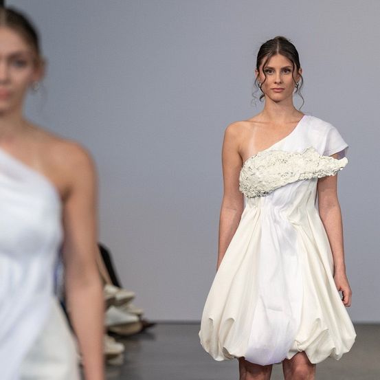 Eine Frau in einem weißen Kleid mit einem einzigartigen Einschulter-Design läuft auf einem Laufsteg, mit verschwommenen Bildern anderer Models und Schuhen im Hintergrund.