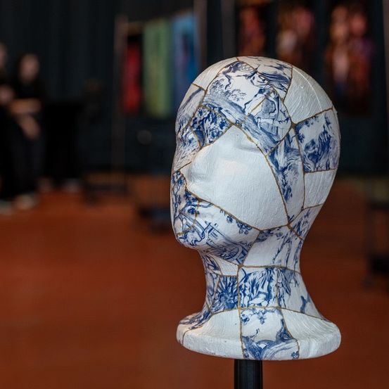 Ein weißer Mannequinkopf mit blauen und weißen Mustern, ausgestellt auf einem Ständer. Im Hintergrund sitzen Menschen auf dem Boden.