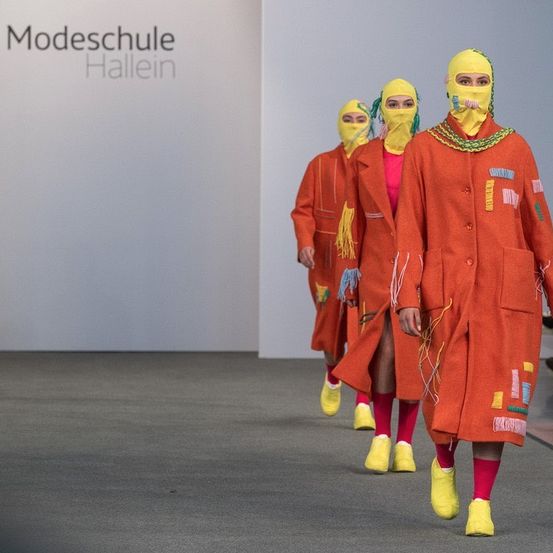 Drei Models in orangefarbenen Mänteln mit gelben Masken und Schuhen laufen auf einem Laufsteg. Dahinter beobachtet ein Publikum von den Tribünen. Ein weißer Hintergrund mit Text steht hinter den Models.