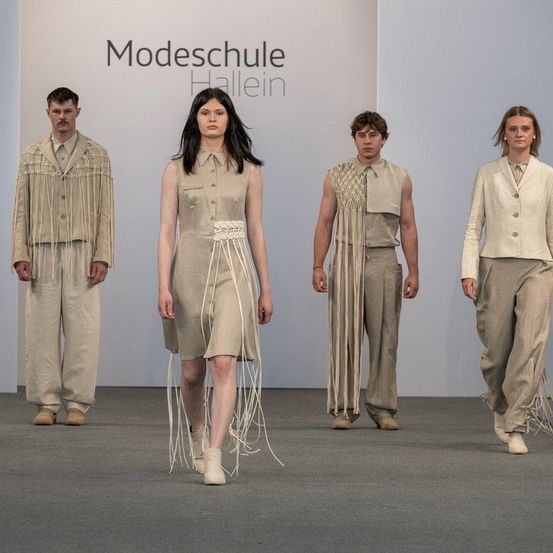 Fünf Personen laufen auf einem Laufsteg für eine Modenschau, jede trägt einzigartige beige Kleidung. Ein weißer Hintergrund zeigt den Text 'Modeschule Hallein'.