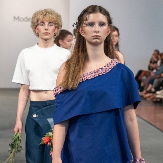 Eine Frau in einem blauen Kleid geht auf dem Laufsteg, gefolgt von einer anderen Frau in Weiß. Dahinter beobachtet ein Publikum die Modenschau.