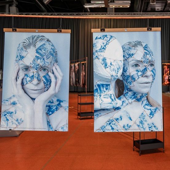 Drei große Poster eines Frauengesichts, das aus blau-weißen Porzellanscherben besteht, hängen auf einem roten Teppich. Das mittlere Poster hat einen weißen Hintergrund.