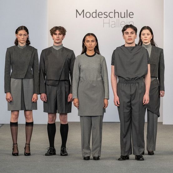 Sechs Models stehen in einer Reihe vor einem weißen Hintergrund mit 'Modeschule Hallein'. Sie tragen graue Anzüge, hohe Absätze und schwarze Socken.
