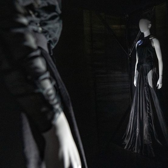 Ein Mannequin in einem schwarzen Kleid ist in einem dunklen Raum ausgestellt. Das Kleid hat einen Schlitz an der Seite. Eine Hand, möglicherweise ein Kleid haltend, ist links.