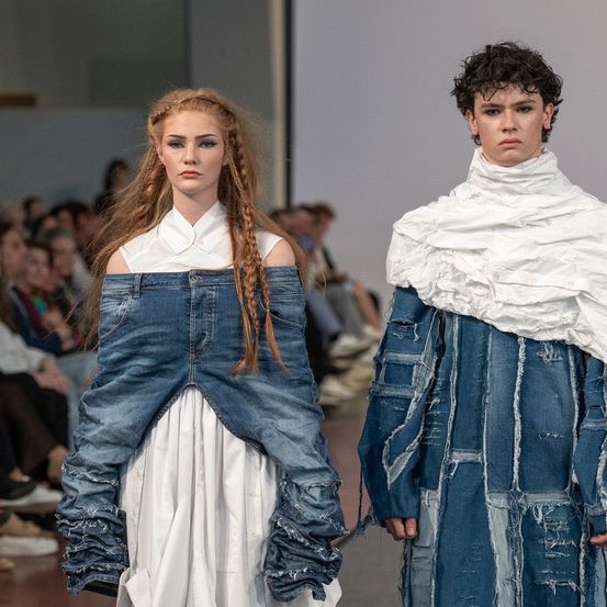 Zwei Models laufen auf dem Laufsteg, eines mit geflochtenen Haaren und einem Jeanskleid, das andere in einem gerüschten weißen und Jeans-Outfit. Dahinter beobachtet ein Publikum.