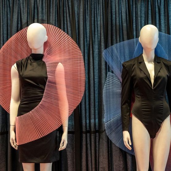 Zwei Mannequins in schwarzen Outfits mit strukturierten Schultern stehen vor blauen Vorhängen. Das linke trägt ein ärmelloses Kleid mit einem fächerartigen roten Kragen. Das rechte trägt ein schwarzes Bodysuit mit Blazer.
