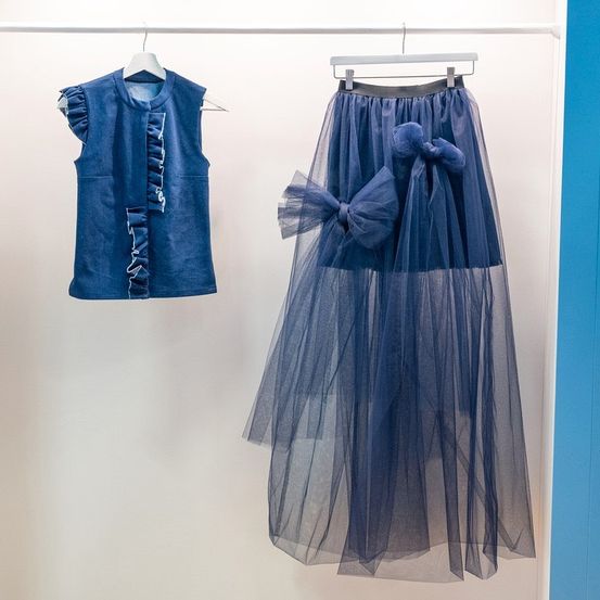 Zwei blaue Kleidungsstücke hängen an einer weißen Wand. Das linke ist ärmellos mit Rüschen und das rechte ist ein Tüllrock mit einer Schleife.