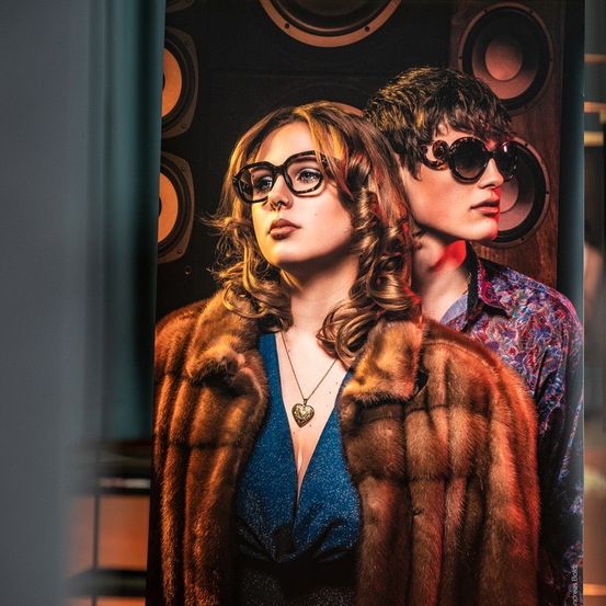 Zwei Menschen stehen nebeneinander. Die Frau trägt einen Pelzmantel und der Mann eine Sonnenbrille. Hinter ihnen befindet sich eine Holzwand mit Lautsprechern.