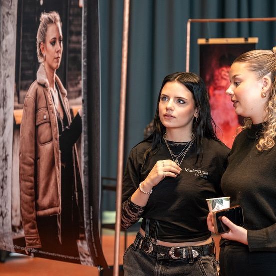 Zwei Frauen stehen vor einem Modeposter einer Frau in einer braunen Jacke. Eine trägt ein schwarzes Shirt mit einer Halskette und hält einen Becher, die andere ein Handy.