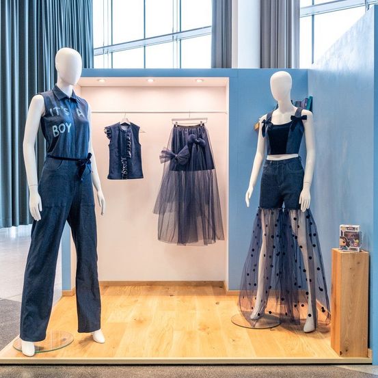 Zwei Mannequins in einem Bekleidungsgeschäft präsentieren verschiedene Denim-Outfits. Das linke Mannequin trägt eine blaue Denim-Jumpsuit mit einem Slogan, während das rechte Mannequin einen Denim-Rock mit Punkten zeigt.