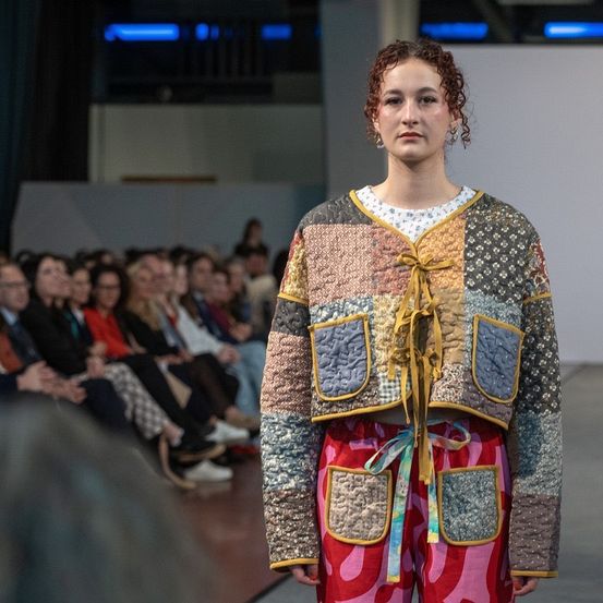 Eine Modenschau zeigt eine Frau in einer bunten Patchwork-Jacke mit gelben Schnüren und rosa Hosen, die über den Laufsteg geht, während ein Publikum zuschaut.