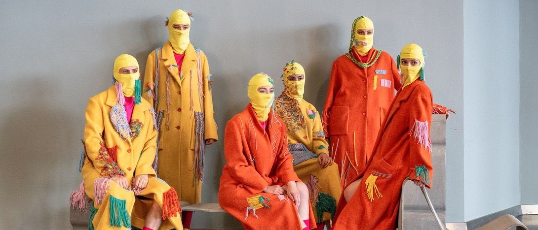 Sieben Personen in passenden gelben Mänteln und Masken mit Fransen sitzen zusammen. Sie tragen gelbe Stiefel und rosa Socken. Der Hintergrund ist eine schlichte graue Wand.