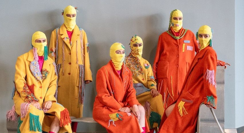 Sieben Personen in passenden gelben Mänteln und Masken mit Fransen sitzen zusammen. Sie tragen gelbe Stiefel und rosa Socken. Der Hintergrund ist eine schlichte graue Wand.