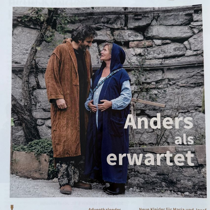 Ein Mann und eine Frau stehen dicht beieinander vor einer Steinmauer. Die Frau trägt einen blauen Umhang und der Mann einen braunen Umhang. Sie schauen sich in die Augen. Der Text lautet 'Anders als erwartet'.