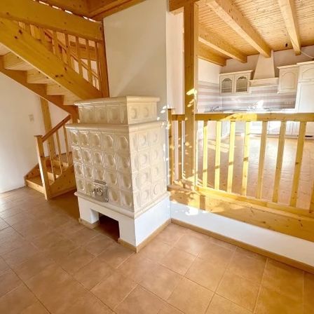 Ein gemütlicher Wohnbereich mit Holzboden, einer Treppe und einem Holzgeländer. Rechts ist eine Küche mit weißen Schränken und einer gelben Arbeitsplatte zu sehen.
