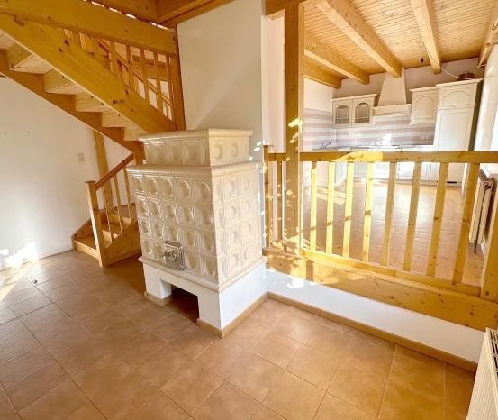 Ein gemütlicher Wohnbereich mit Holzboden, einer Treppe und einem Holzgeländer. Rechts ist eine Küche mit weißen Schränken und einer gelben Arbeitsplatte zu sehen.