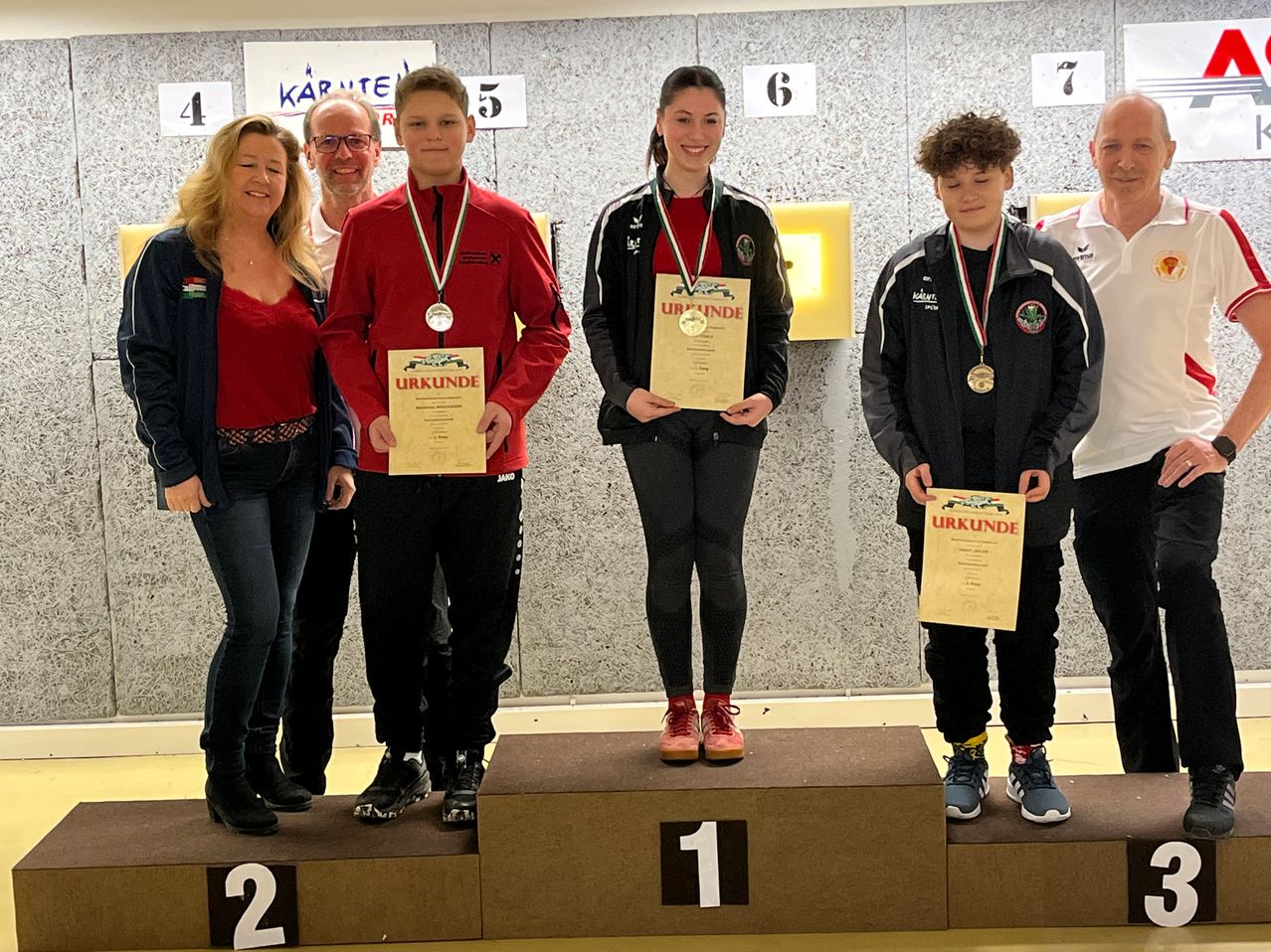 Vier Personen stehen auf einem Podest mit Medaillen um den Hals. Sie halten Urkunden in den Händen, und das Podest hat die Nummern 1, 2 und 3. Dahinter befindet sich eine Wand mit den Nummern 4, 5, 6 und 7.