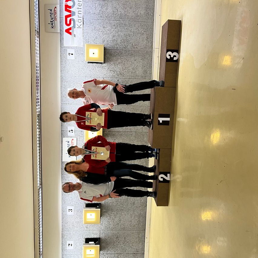 Vier Personen stehen auf einem Podium mit den Nummern 1, 2 und 3. Sie halten Medaillen und scheinen zu feiern. Dahinter befinden sich Schilder mit Nummern und Text.