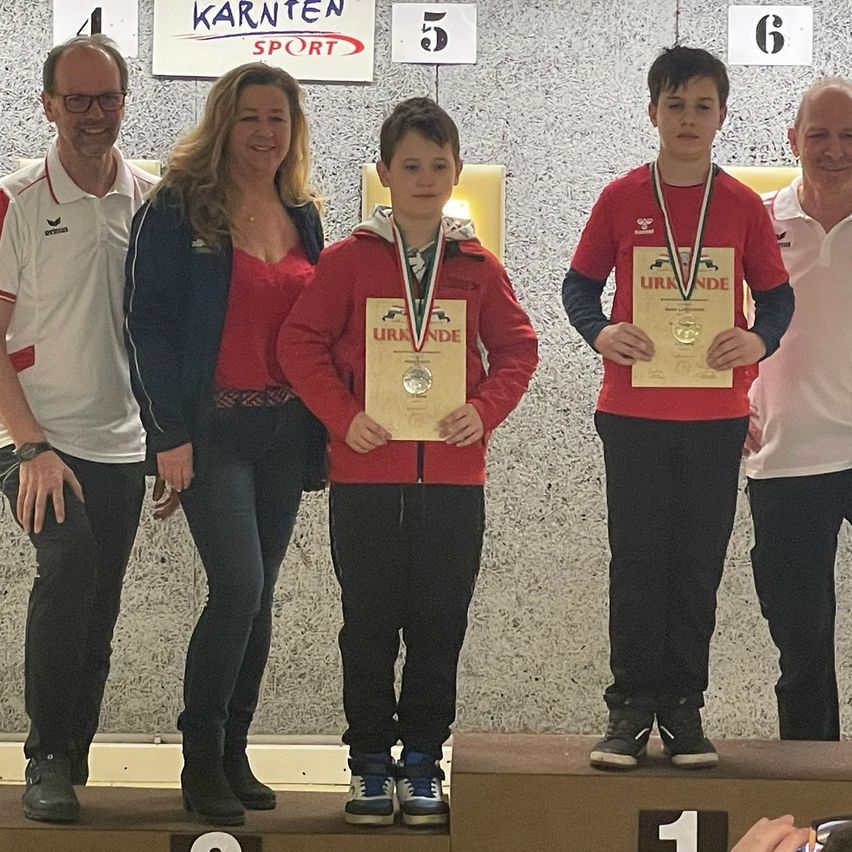 Zwei Jungen stehen auf dem Podium und halten Medaillen. Sie tragen rote Jacken und schwarze Hosen. Dahinter stehen zwei Erwachsene, einer trägt ein weißes Hemd und eine Brille, der andere ein rotes Oberteil und blaue Jeans. Dahinter ist eine graue Wand mit den weißen Nummern 5 und 6.