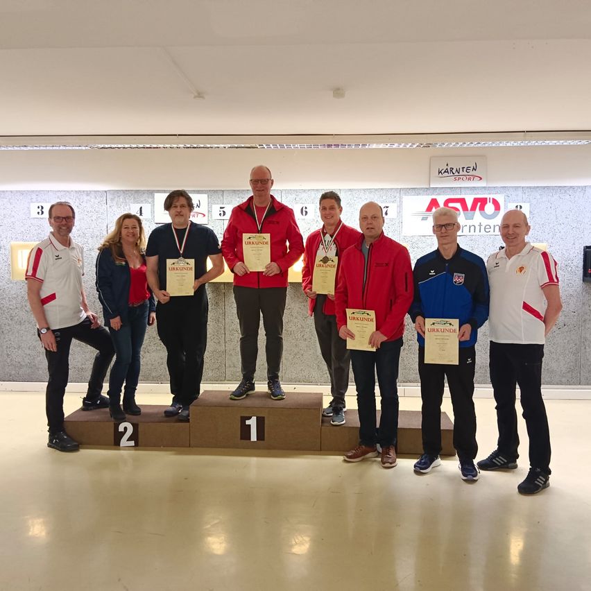 Acht Personen stehen auf einem Podest in einem Raum. Der Mann in der Mitte trägt eine rote Jacke. Das Podest hat die Zahlen 1, 2 und 3. Dahinter befindet sich eine Wand mit den Nummern 1 bis 7 und den Texten 'ASVO' und 'Kärnten Sport'.