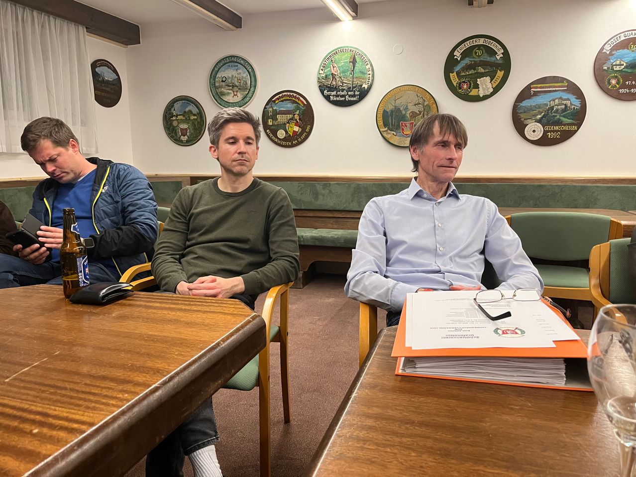 Zwei Männer sitzen in Stühlen gegenüber, mit einem Tisch dazwischen. Sie scheinen sich in einem Meeting zu befinden. Der Mann rechts hat einen Stapel Papiere und eine Brille auf dem Tisch. Hinter ihnen hängen mehrere gerahmte Plaketten an der Wand.