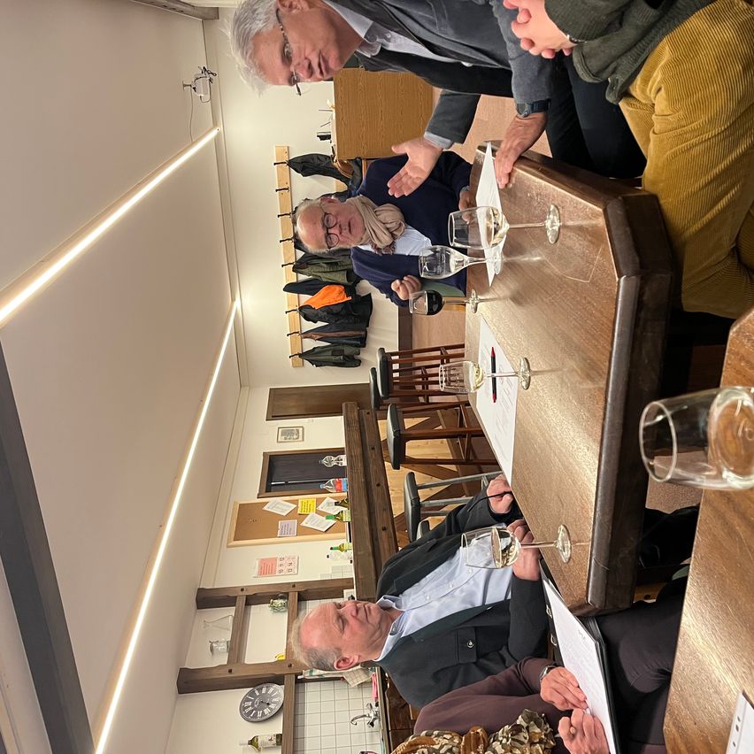 Mehrere Männer sitzen um einen Holztisch herum und führen eine Diskussion. Gläser mit Wein stehen auf dem Tisch, und die Umgebung scheint ein zwangloses Treffen zu sein. Der Raum ist gut beleuchtet mit weißen Wänden und einer Decke.