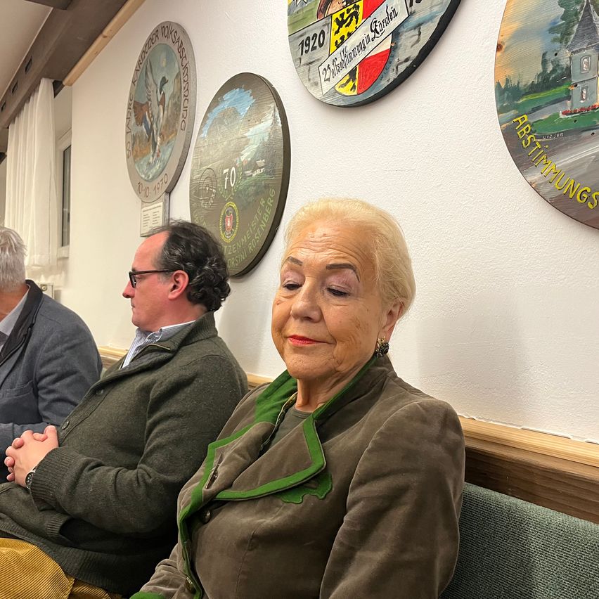 Drei Menschen sitzen in einem Raum. Eine Frau mit weißen Haaren sitzt in der Mitte. Zwei Männer sitzen auf beiden Seiten von ihr. Eine weiße Wand ist hinter ihnen mit mehreren Rahmen, die daran hängen.