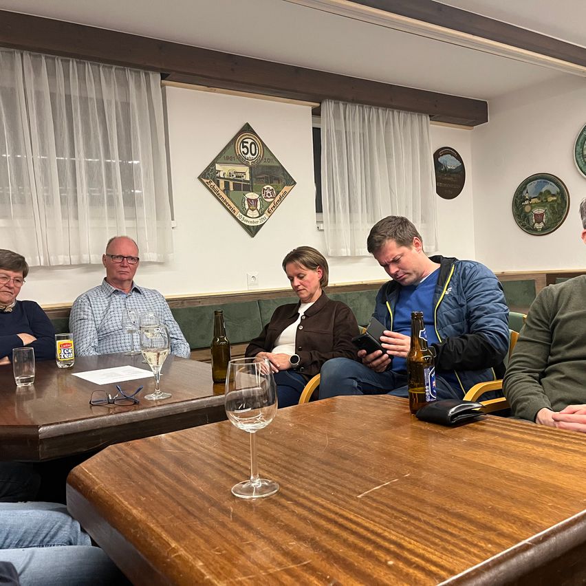 Fünf Menschen sitzen um einen Holztisch herum, drei Männer und zwei Frauen. Der Mann in der Mitte hat den Kopf gesenkt und hält ein Telefon. Der Tisch ist mit Gläsern, Flaschen und Papieren übersät. Hinter ihnen hat die Wand Vorhänge und dekorative Teller.