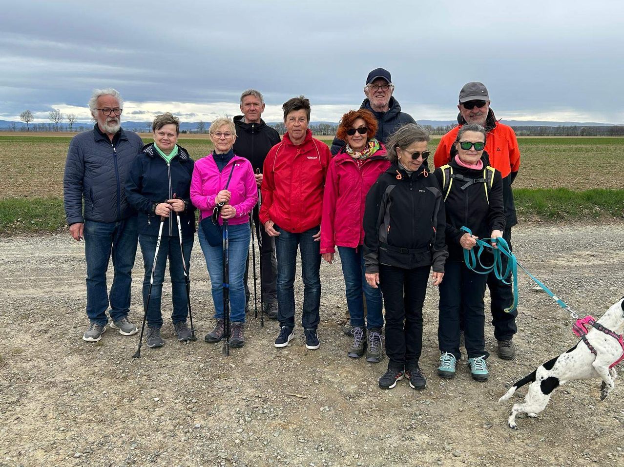 Eine Gruppe von Menschen in Outdoor-Kleidung, die Trekkingstöcke halten, posiert für ein Foto auf einem Erdweg mit einem bewölkten Himmel im Hintergrund.