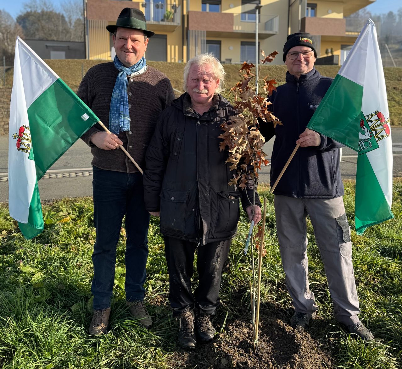 Drei Männer stehen vor einem kleinen Baum und halten Flaggen mit grün-weißem Design. Sie lächeln und scheinen für ein Foto zu posieren. Hinter ihnen befindet sich ein Gebäude mit gelber Außenfassade.