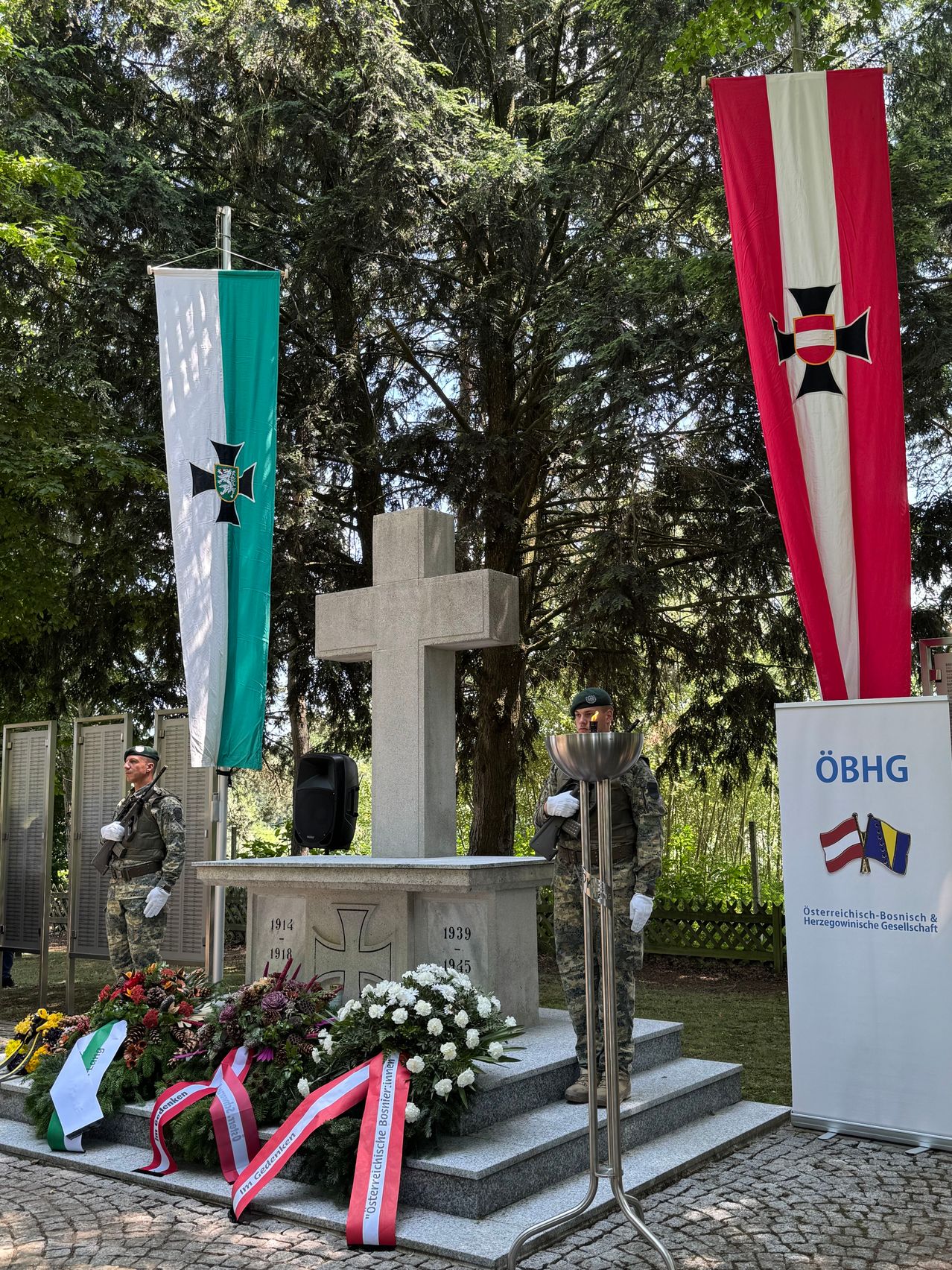 Zwei Soldaten stehen vor einem Gedenkkreuz mit Blumen an seiner Basis. Hinter ihnen zeigt ein Fahnenmast zwei Flaggen, eine mit einem schwarzen Kreuz auf weißem Hintergrund und die andere mit einem roten und weißen Design. Das Denkmal hat Tafeln mit den Jahren 1912-1918 und 1939-1945.