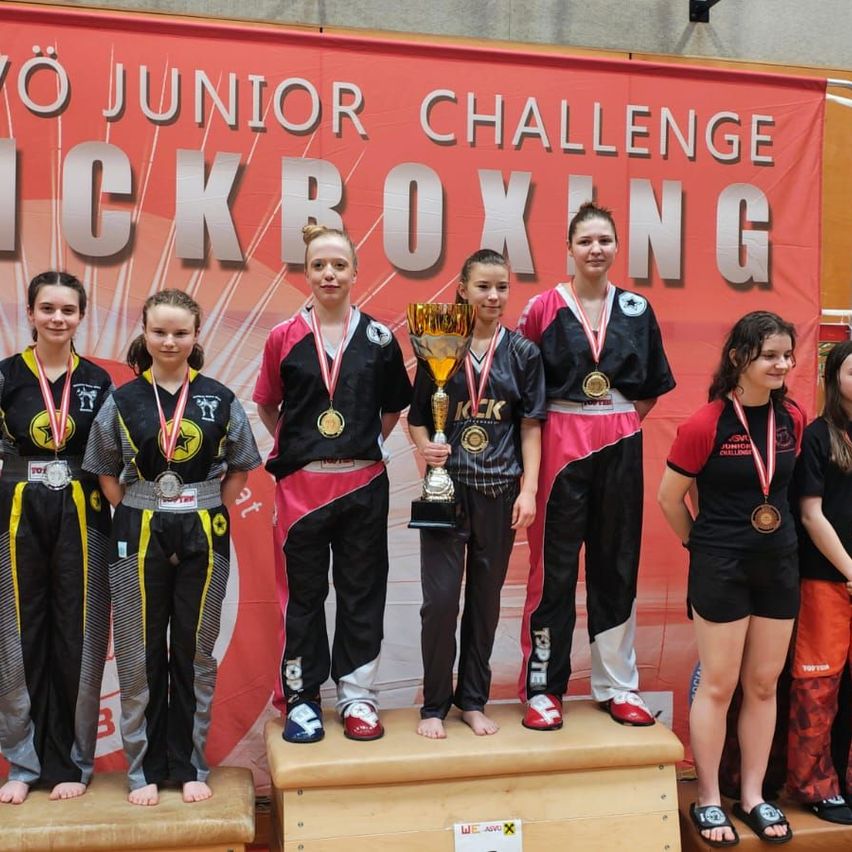 Fünf junge Frauen stehen auf einer Bühne, eine von ihnen hält einen Pokal in der Hand, trägt Medaillen und hinter ihnen befindet sich ein Banner mit der Aufschrift 'Junior Challenge Kickboxing'.
