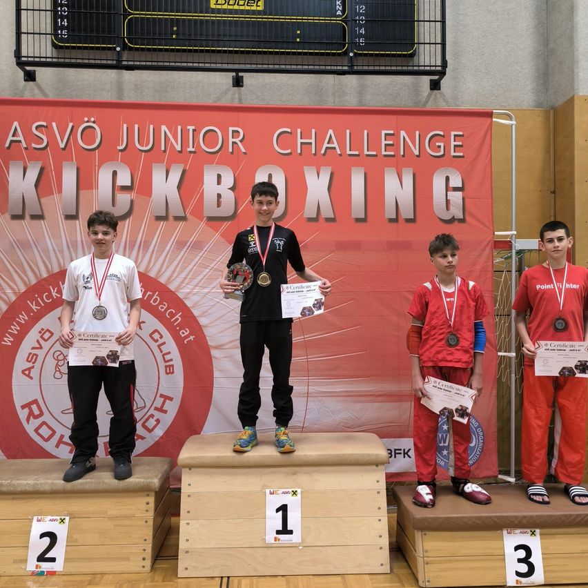 Vier Jungen stehen auf der Bühne und halten ihre Medaillen und Urkunden. Der Junge in der Mitte trägt ein schwarzes T-Shirt und Turnschuhe. Der Junge auf der linken Seite trägt ein weißes T-Shirt und Turnschuhe. Der Junge auf der rechten Seite trägt ein rotes T-Shirt und Turnschuhe.