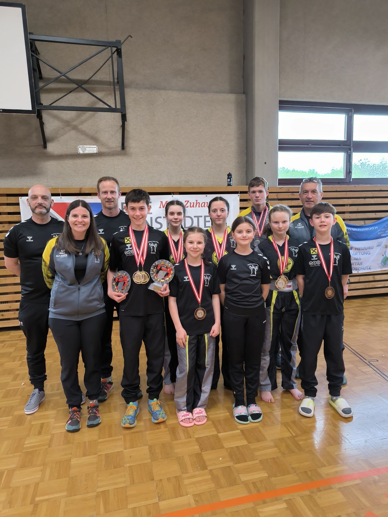 Eine Gruppe junger Athleten, die schwarze T-Shirts, schwarze Hosen und Turnschuhe tragen, steht zusammen auf einem Holzboden. Sie halten Medaillen und lächeln, mit einer Frau in einer grauen Jacke, die vor ihnen steht. Dahinter stehen Menschen und ein Banner.