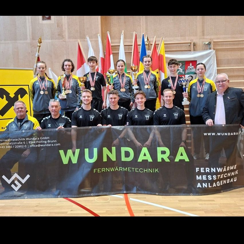 A group of young athletes stand together, holding trophies and medals. They wear sports uniforms and stand in front of a banner with the name Wundara. Behind them, flags are displayed.