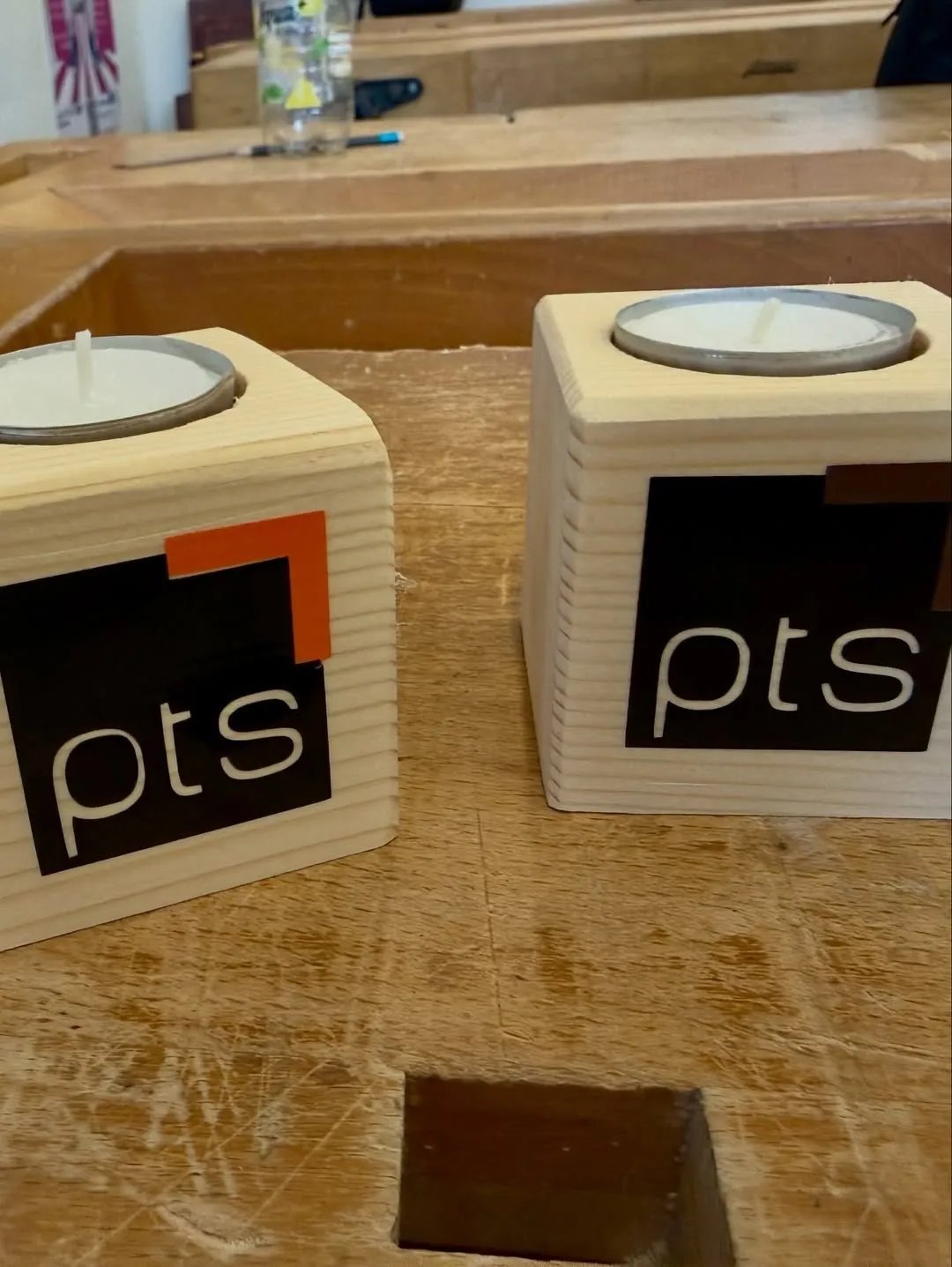 Zwei hölzerne Kerzenhalter mit dem Label 'pts' stehen auf einer Holzoberfläche. Jeder Halter hat eine Kerze darin, einer mit einem orangefarbenen Akzent.